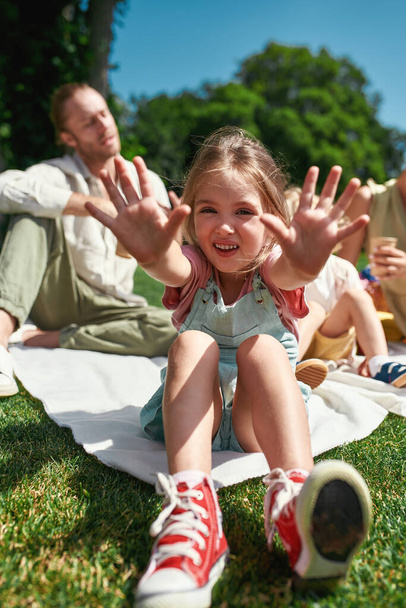 Cute little girl reaching hands to camera while relaxing with family outdoors, having picnic in nature on a summer day - Foto, imagen