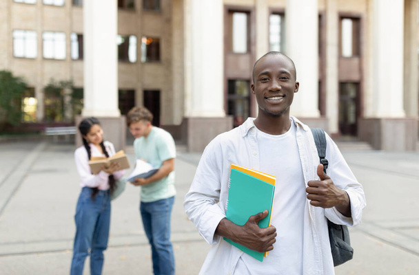 Щасливий афроамериканець, який стоїть на вулиці з однокласниками в університетському містечку, подає і посміхається фотоапарату. - Фото, зображення