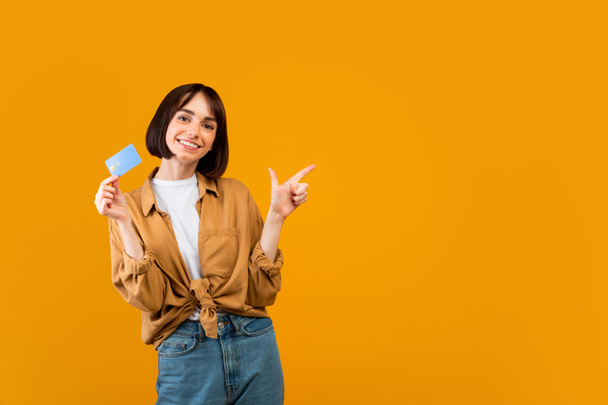 Innoissaan nainen luottokortilla osoittaa sivuun tyhjään tilaan keltainen studio tausta, panoraama - Valokuva, kuva