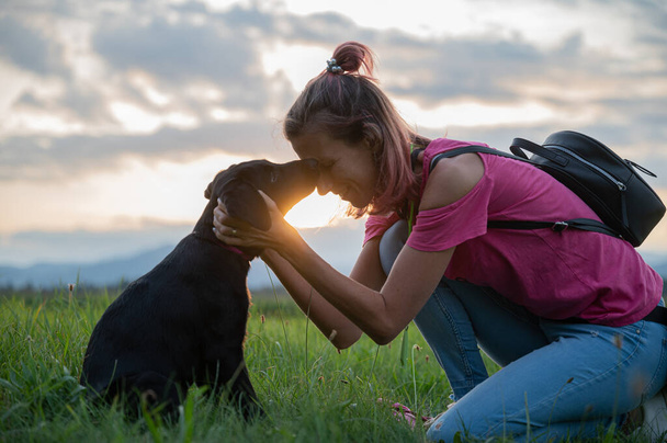 Joven cachorro labrador besando a su dueña mientras se arrodilla ante ella en un hermoso prado verde con el sol poniente brillando entre ellos. - Foto, imagen