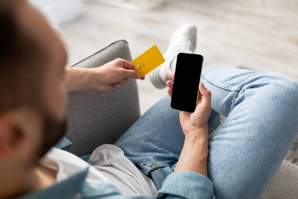Unerkennbarer Millennial-Mann mit Kreditkarte und Handy von zu Hause aus online einkaufen, App-Attrappe oder Webseite - Foto, Bild
