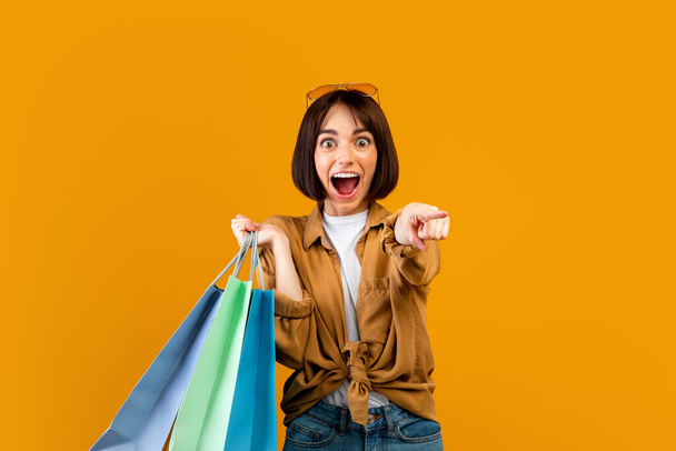 Hey, you. Excited woman holding colorful shopping paper bags and pointing at camera over orange studio background - Фото, изображение