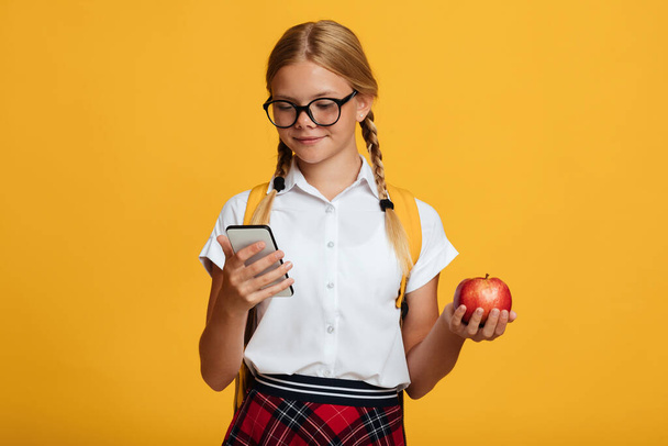 Happy young blonde girl pupil with pigtails and backpack in glasses typing on phone, holds red apple - Fotó, kép