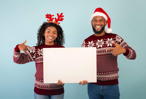 Glückliches afrikanisch-amerikanisches Paar zeigt auf leeres Weihnachtsposter, posiert vor blauem Hintergrund, Kopierraum - Foto, Bild