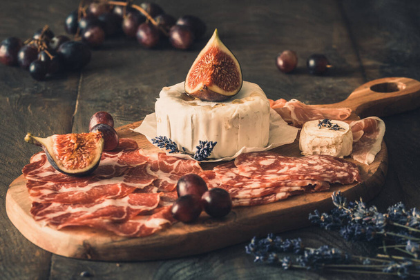Diverses spécialités de saucisses fumées et un fromage à pâte molle sur une planche de bois, décorée de figues et de raisins - Photo, image