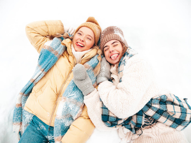 Two young beautiful smiling hipster female in trendy warm clothes and scarfs.Carefree women posing in the street in park. Positive pure models lying in snow. Enjoying winter moments. Xmas concept - Фото, изображение
