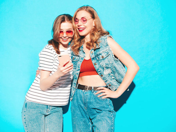 Two young beautiful smiling hipster female in trendy summer clothes. Sexy carefree women posing near blue wall in studio.Trendy and positive models looking at smartphone screen. Using phone apps - Φωτογραφία, εικόνα