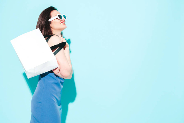 High fashion portrait of young beautiful brunette woman wearing nice blue summer dress.Sexy trendy model posing near blue wall in studio.Fashionable female in sunglasses. Holding shopping bag - 写真・画像