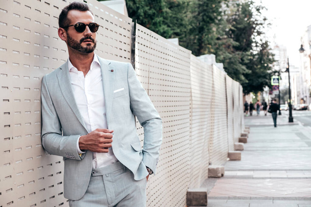 Portrait of handsome confident stylish hipster lambersexual model.Sexy modern man dressed in elegant white suit. Fashion male posing in the street background near fence at sunset. In sunglasses - Φωτογραφία, εικόνα