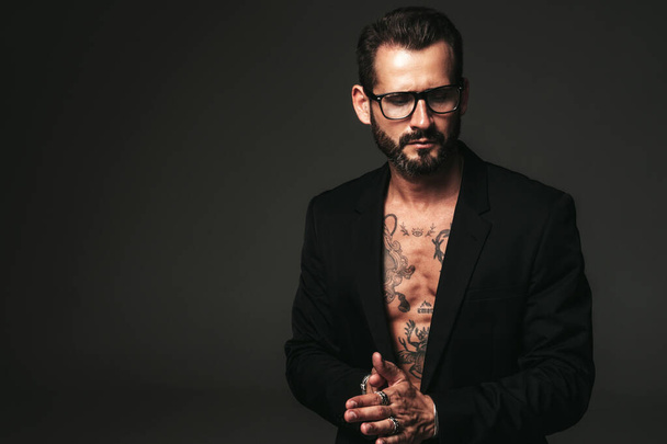 Portrait of handsome confident stylish hipster lambersexual model. Sexy modern man dressed in black jacket. Naked torso with tattoos.Fashion male posing in studio on dark background. In spectacles - Photo, Image
