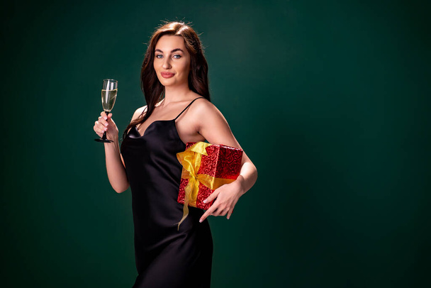 Žena v černých šatech drží dárkovou červenou krabici a sklenici šampaňského. Čas na párty. Koncept dovolené - Fotografie, Obrázek