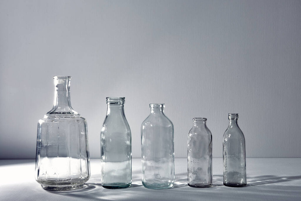 A five different empty glasses bottles with copy space. Graphic still life with light and shadow in gray light. The concept of containers, recyclable materials or alcoholism. Creative vintage vase. - Фото, зображення