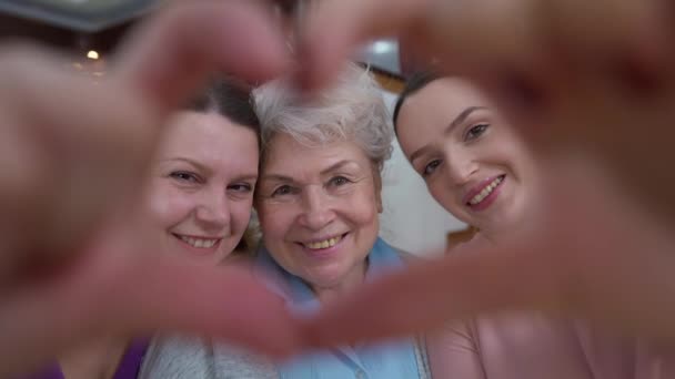 Close-up female hands in heart shape with smiling adult senior and young women looking at camera smiling. Portrait of positive happy Caucasian multigenerational family posing indoors at home. - Footage, Video