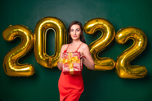 Smiling brunette woman in red dress raises the gift red box up. NEW YEAR 2022 gold air balloons.  - Φωτογραφία, εικόνα