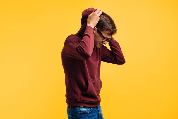 young guy with glasses and red sweater puts on hood over yellow background, autumnal concept - Фото, изображение