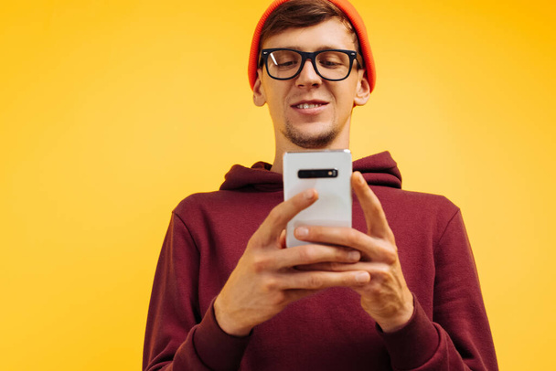 handsome young guy in an orange hat with glasses and a red sweater is typing a message on the phone, holding the phone in his hands, standing on a yellow background, autumnal concept, 5g - 写真・画像