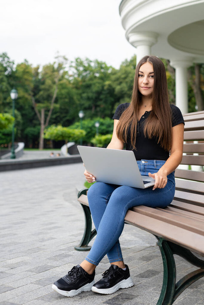 Educación, negocios, tecnología y concepto de Internet: chica morena europea vestida con ropa casual sentada en un banco del parque y sosteniendo una computadora portátil. - Foto, Imagen
