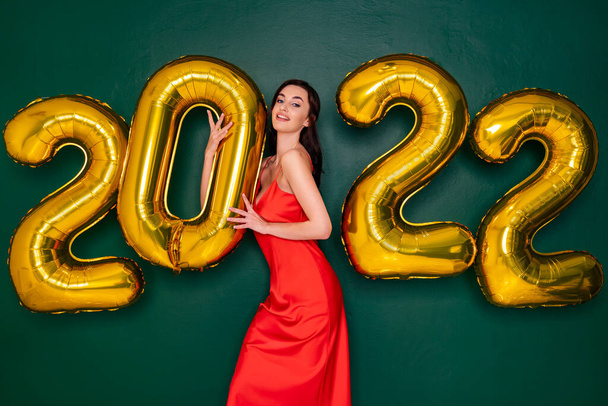 Young happy woman in red dress holding 2022 golden balloons with green backdrop. New Year concept. - Foto, Imagem