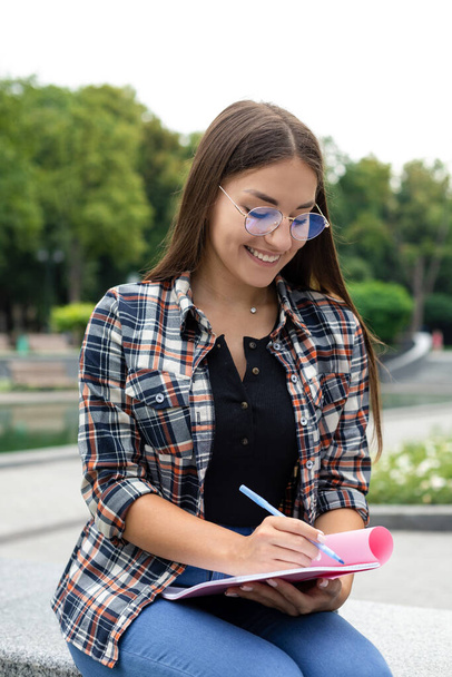 Studiowanie, świeże powietrze, technologia - młoda biała kobieta w koszuli w kratę i okularach siedzi w parku, pisząc notatki w dużym notatniku podczas konferencji internetowej. - Zdjęcie, obraz