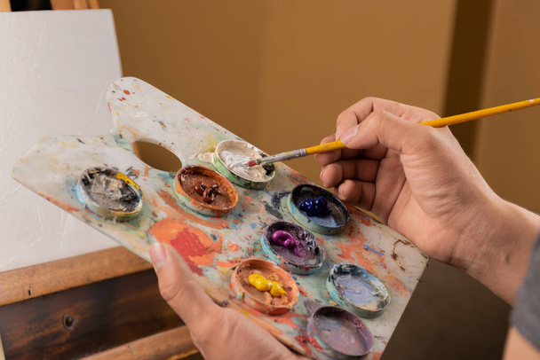 Boya lekeli genç bir elin boya fırçası, paletin renklerini karıştıran sanatçı, iş aletleri, hobi, stüdyoda yaratıcı yaşam tarzı. - Fotoğraf, Görsel