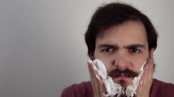 Brutal man is putting mousse on cheeks and shaving beard by the axe frowning his eyebrows - Footage, Video