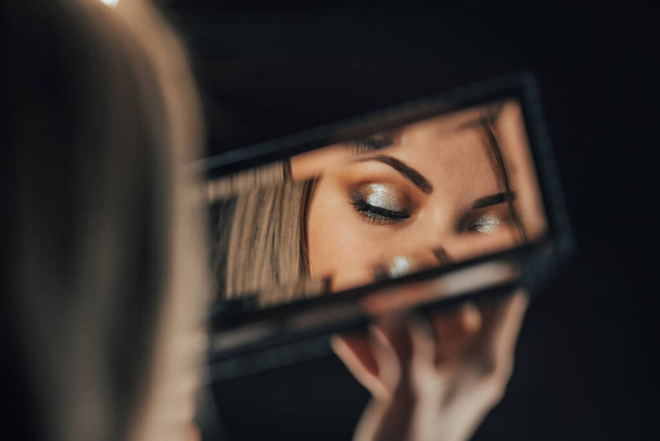 Reflection in mirror of womans eyes with shinning make-up. Young lady looks at herself. Makeup at night getting ready before going to party. - Photo, image