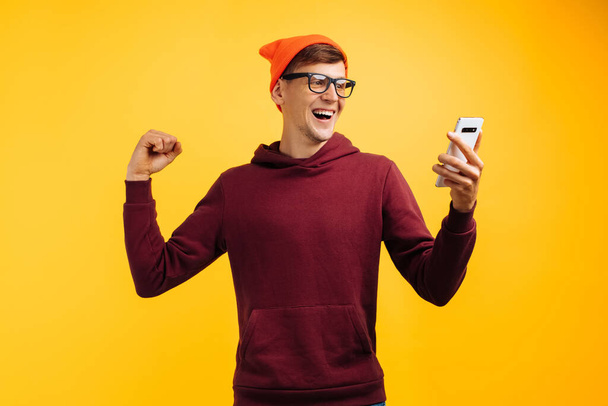 stylish young guy in an orange hat with glasses and a red sweater guy rejoices at a message on the phone, demonstrates success raising his hands up, autumn mood - Photo, Image