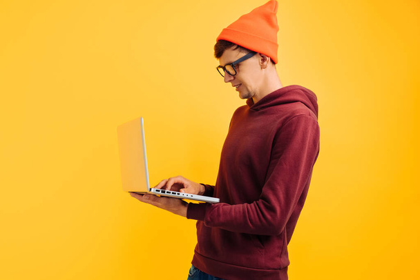 young handsome guy with typing on a laptop, in an orange hat with glasses and a red sweater, on a yellow background, high-speed internet, 5g - Photo, Image