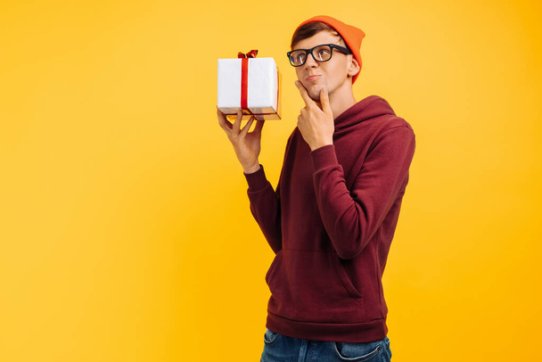 handsome young guy in an orange hat with glasses and a red sweater, thinking what is inside his gift, festive mood, autumn concept, hands with a white cork and a red bow - Foto, afbeelding