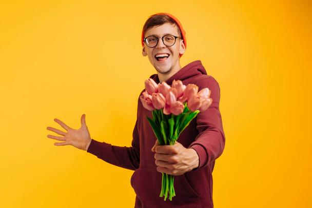 a handsome man with a bouquet of flowers with delight gives flowers wrapped in pink paper, a man in an orange hat and a red sweater with flowers in his hands, on a yellow background - Photo, image
