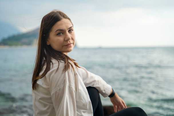 Young brunette in white shirt is sitting on rocky seacoast. - Zdjęcie, obraz