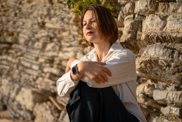 Attractive young woman in fashionable white shirt with haughty look is sitting by a rock hugging her knees. She reflects on the past and makes plans for the future. Place for meditation and reflection - Fotografie, Obrázek