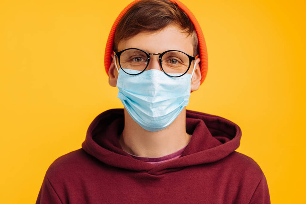 handsome man in a protective mask. in glasses and an orange hat and a red sweatshirt, smiling at the camera, on a yellow background, happy vaccinated man, close-up - Foto, Imagem