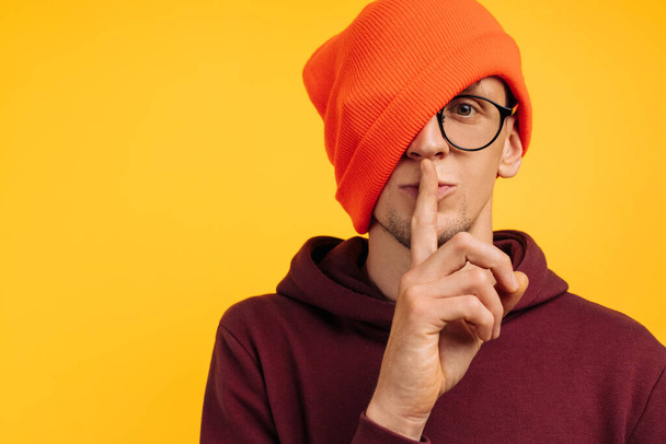 handsome man holds his finger near his lips asks for silence, in an orange hat and a red sweater and glasses, on a yellow background - Fotó, kép