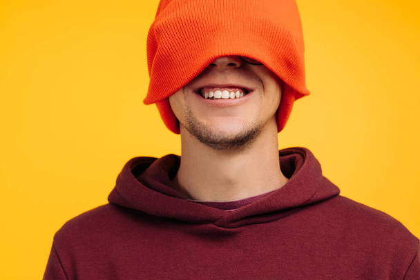 ハンサムな男は彼の顔の床にオレンジの帽子をかぶります,陽気な肯定的な笑顔の男のポーター,黄色の背景に,概念:秋や冬の断熱材,寒い天候のための服 - 写真・画像