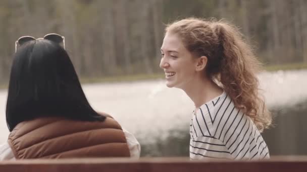 Langzame opname van twee jonge vrolijke vriendinnen die op een houten bankje bij het meer zitten, bierflesjes roosteren en praten, samen genieten van de natuur - Video