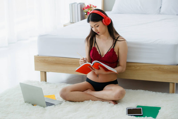 Όμορφη νεαρή άτακτη σέξι κορίτσι στο κόκκινο σουτιέν καθίσει πίσω από λευκό κρεβάτι τέλος ευτυχώς χαμογελώντας καθώς απολαμβάνει το διάβασμα του βιβλίου και τη μελέτη των μέσων διάλεξη από το laptop, tablet, και κόκκινο ακουστικό σε φωτεινό πρωινό - Φωτογραφία, εικόνα