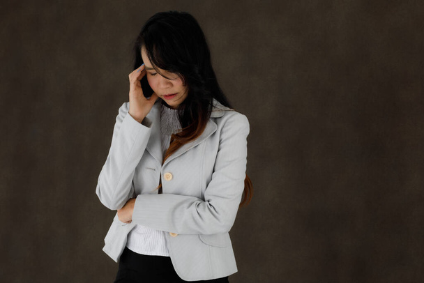 Серьёзная задумчивая молодая азиатская бизнесвумен в шикарном наряде трогает лоб и задумчиво закрывает глаза на темно-сером фоне с пустым пространством - Фото, изображение