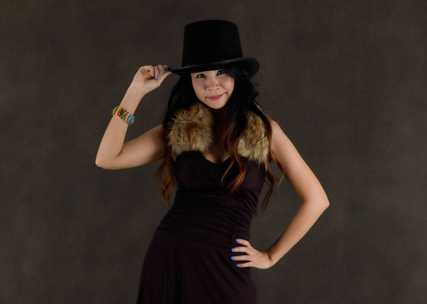 Счастливая молодая азиатка в элегантном черном платье с меховым воротником и стильной шляпой ярко улыбается во время вечеринки на черном фоне - Фото, изображение