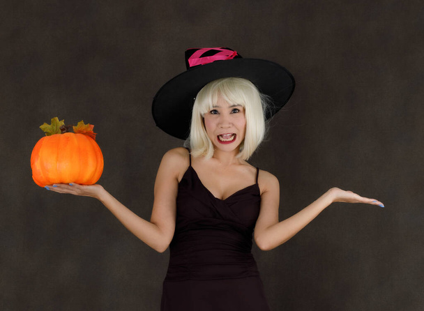 Young Asian female in blond wig and witch hat with Halloween pumpkin laughing happily during party on black background - 写真・画像