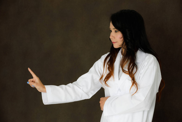 Νεαρή ασιατική γυναίκα γιατρός με στηθοσκόπιο θέτουν μοιάζουν με αγγίζοντας αόρατο ή εικονικό κουμπί πραγματικότητα σε σκούρο γκρι φόντο - Φωτογραφία, εικόνα
