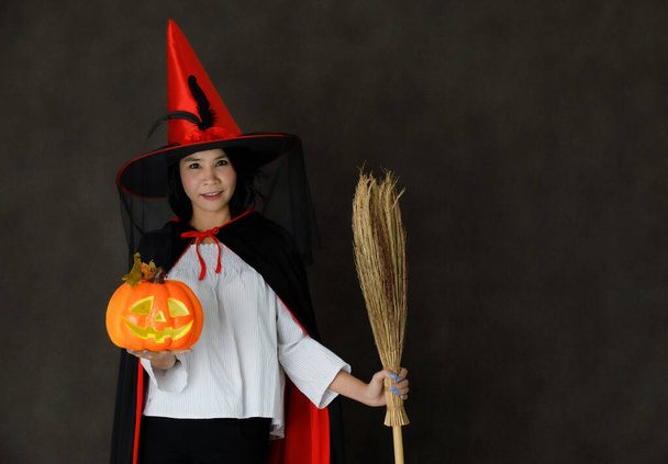 Charmante glimlachende jonge Aziatische vrouw in heks kostuum met Halloween pompoen en bezem kijken naar camera tegen donker grijze achtergrond - Foto, afbeelding