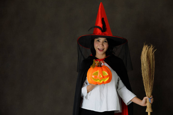 Urocza uśmiechnięta młoda Azjatka w stroju czarownicy z dyni Halloween i miotły patrząc na kamerę na ciemnym szarym tle - Zdjęcie, obraz