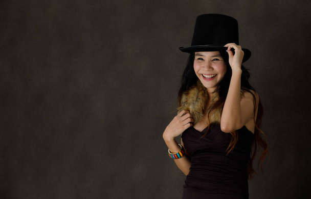 Jovem mulher asiática feliz em vestido preto elegante com gola de pele e chapéu elegante sorrindo brilhantemente durante o partido contra fundo preto - Foto, Imagem