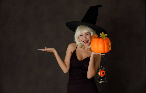 Junge Asiatin mit blonder Perücke und Hexenhut mit Halloween-Kürbislaterne lacht fröhlich bei Party auf schwarzem Hintergrund - Foto, Bild