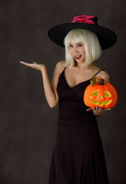 Joven hembra asiática en peluca rubia y sombrero de bruja con calabaza de Halloween riendo felizmente durante la fiesta sobre fondo negro - Foto, Imagen