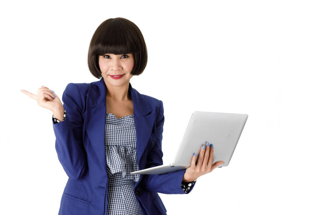 Positive successful young Asian female in elegant blue outfit looking at camera and pointing finger away while working on laptop against white background - Photo, Image