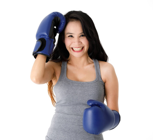 Αποφασισμένη νεαρή ταιριάζει ασιατική γυναίκα σε μπλε γάντια πυγμαχίας στέκεται σε στάση μάχης και κοιτάζοντας κάμερα με χαμόγελο σε λευκό φόντο - Φωτογραφία, εικόνα