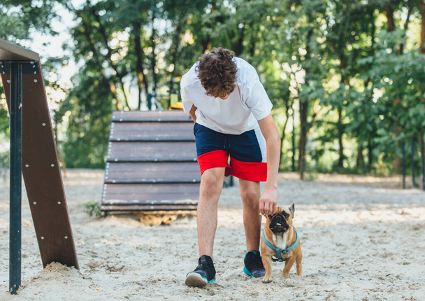 Egy francia bulldog fiú gazdája tanítja a kutyát a parkban. Kutya edzés tinédzser és bulldog a szabadban. Csendélet, házi kedvencek szeretete és gondozása, állatbarátság. - Fotó, kép