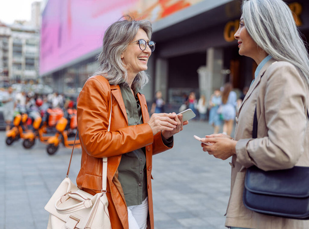 Χαμογελώντας ώριμες γυναίκες και ασιατική κυρία με σύγχρονα κινητά τηλέφωνα μιλούν στο δρόμο της πόλης - Φωτογραφία, εικόνα